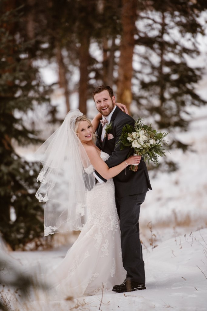 A bride and groom embrace in a snowy forest, surrounded by evergreen trees in Grand County Colorado.