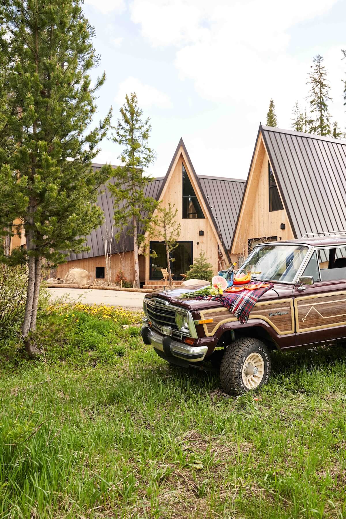 A vintage wood-paneled SUV is parked on a grassy area in front of modern A-frame cabins surrounded by trees in Grand County Colorado.