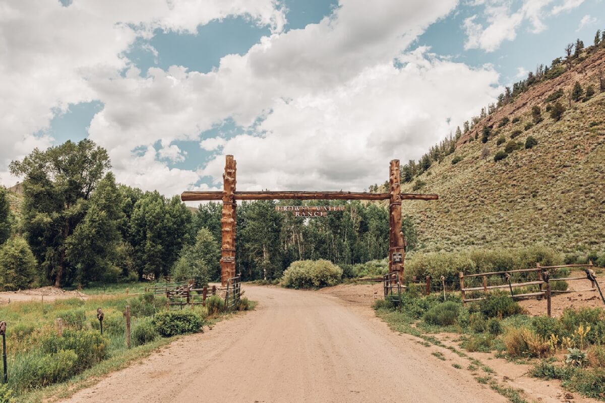 A dirt road winds through a lush, green valley with a rustic wooden archway in the distance in Grand County Colorado.