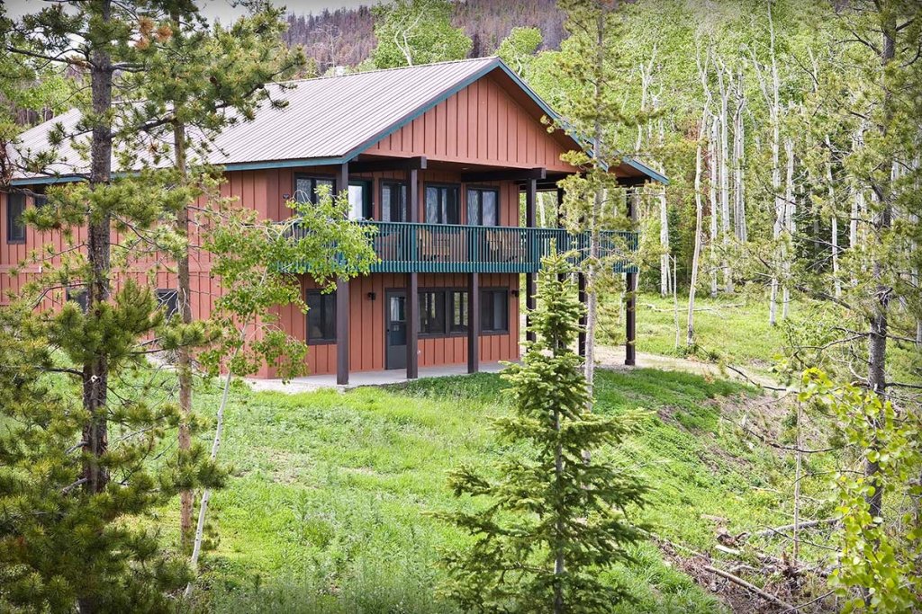 A rustic red cabin with a green balcony is nestled among tall trees in a lush green meadow in Grand County Colorado.