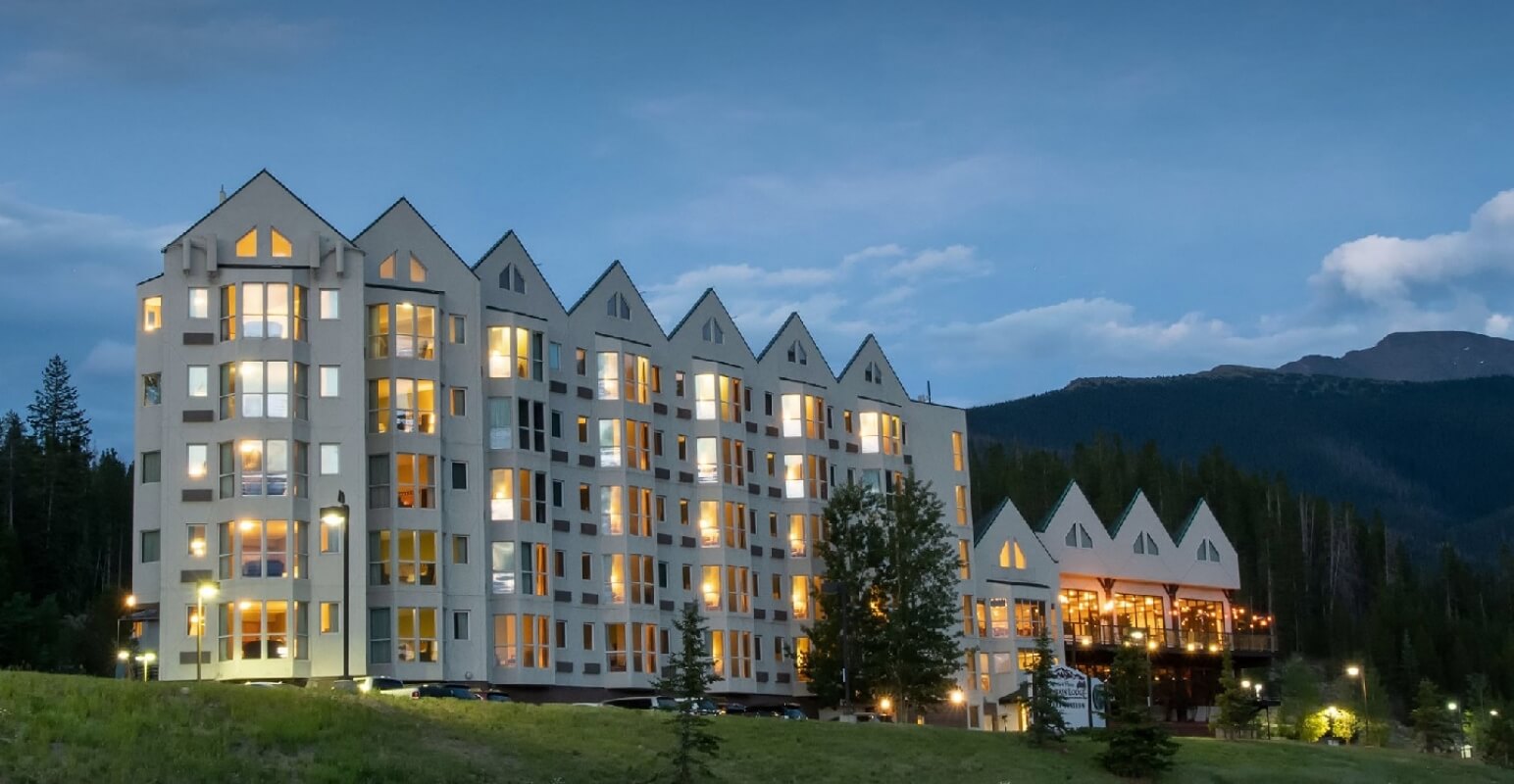 A large, multi-story hotel with a distinctive roofline is nestled among trees at dusk in Grand County Colorado.