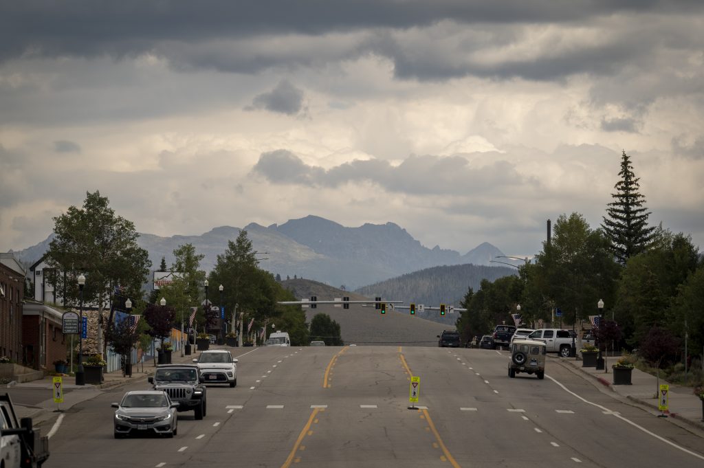 Cars driving along Main Street in Granby with shops, trees, and mountains in the background under cloudy skies.