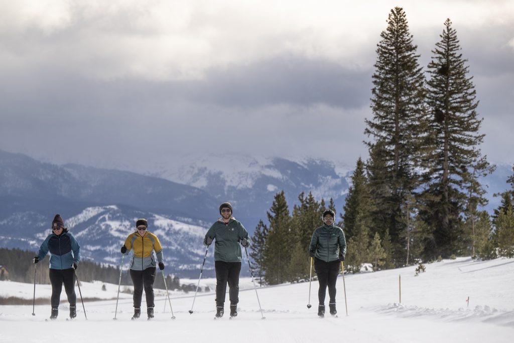 Four people cross-country skiing at YMCA of the Rockies in Grand County during winter, with pine trees and mountains in the background.