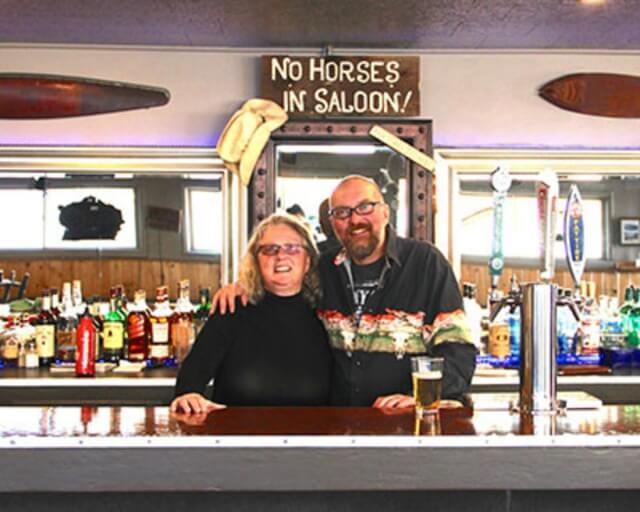 A smiling couple stands at a bar with a 'No Horses in Saloon' sign above them in Grand County Colorado.