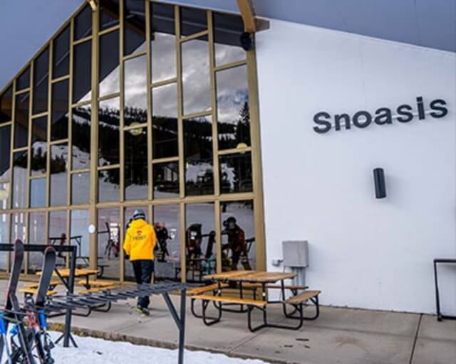 A skier in yellow walks towards a modern ski lodge with large windows and picnic tables outside in Grand County Colorado.