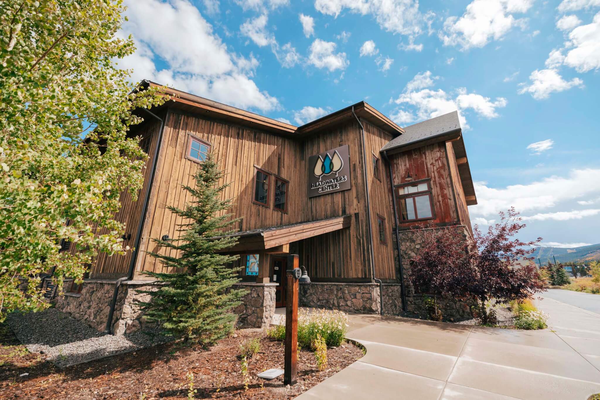 A rustic wooden building with a stone foundation, surrounded by lush greenery and tall trees, stands under a clear blue sky with fluffy white clouds in Grand County Colorado.