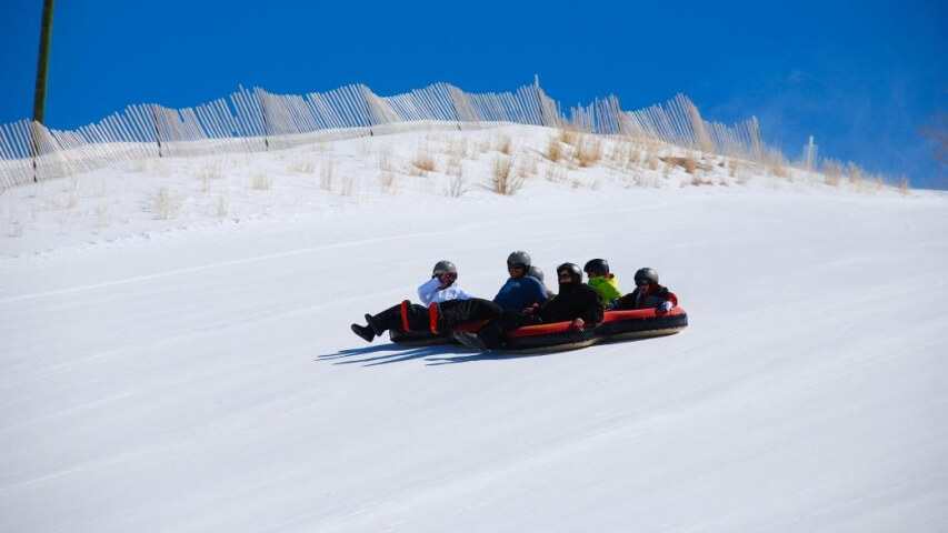 Group of five people snow tubing down a snowy hill, wearing helmets and life jackets in Grand County Colorado.