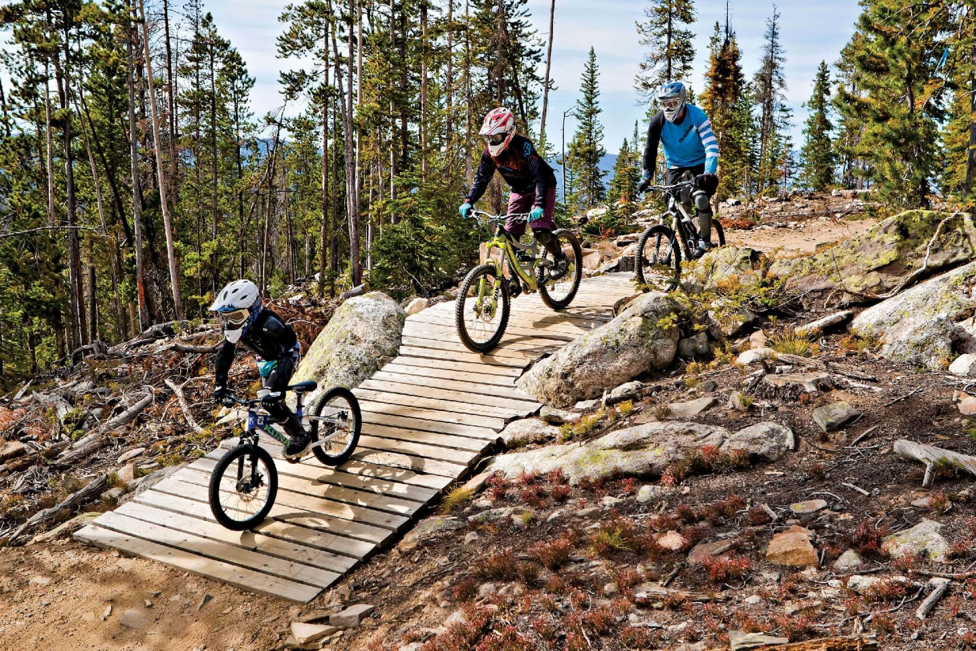 Three mountain bikers, including a child, ride down a wooden ramp in a forest in Grand County Colorado.