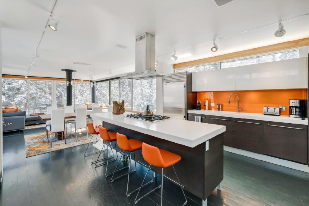 Modern kitchen with orange accents, stainless steel appliances, and large windows overlooking snowy landscape in Grand County Colorado.