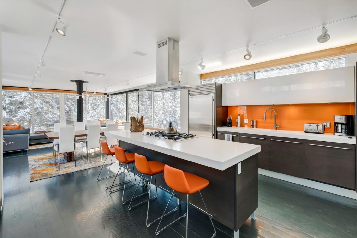 Modern kitchen with orange accents, stainless steel appliances, and large windows overlooking snowy landscape in Grand County Colorado.