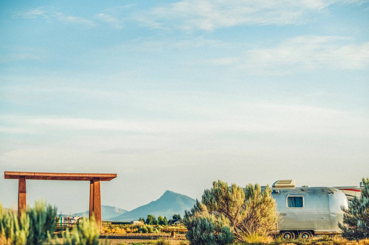 A scenic view of Kremmling, Colorado, featuring an open landscape with sagebrush, a rustic wooden archway, and a vintage Airstream trailer set against distant mountains.