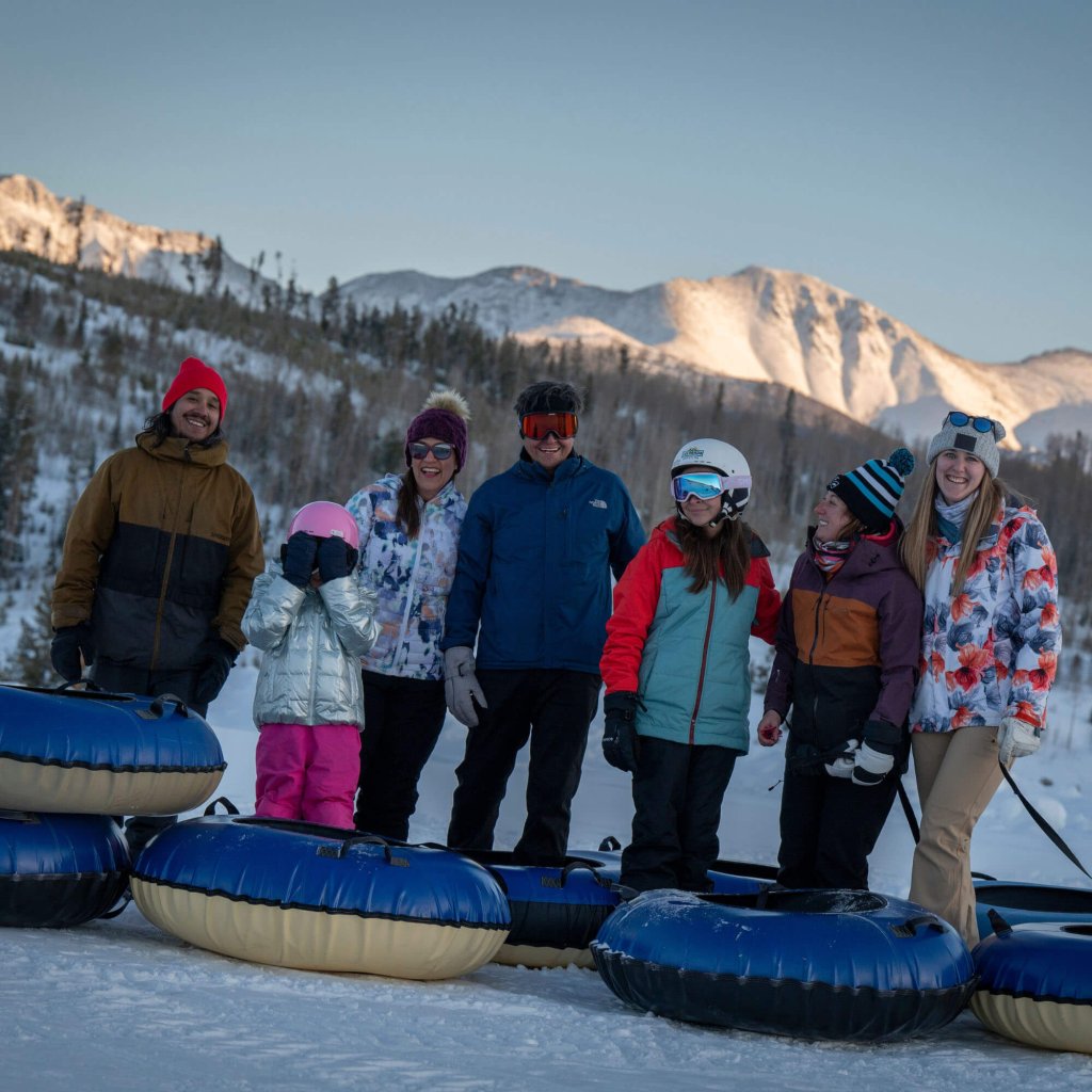 A group of seven people, including a child, are standing in a snowy landscape with blue and yellow inner tubes, ready for a fun day of snow tubing against a backdrop of snow-capped mountains in Grand County Colorado.