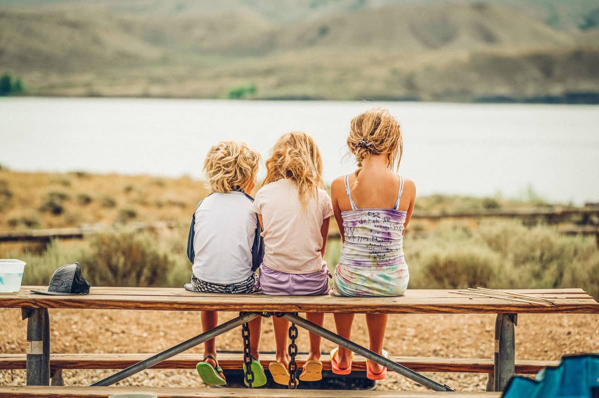 Three kids sitting on a picnic table bench facing the water in Grand County, Colorado.