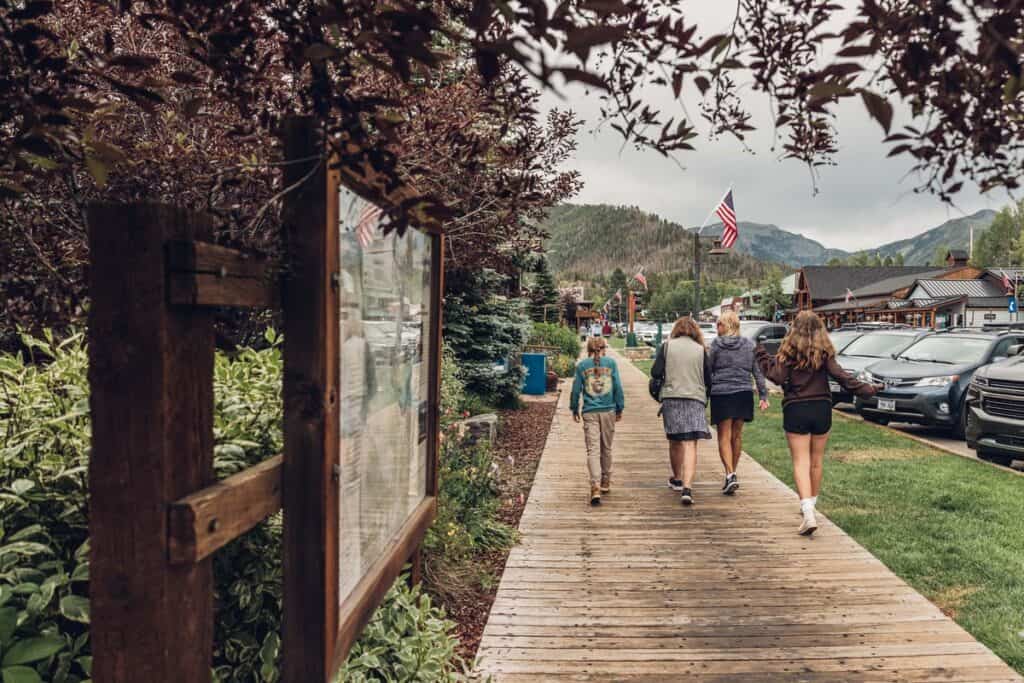 A group of people walk along a wooden boardwalk in a small town with parked cars on one side and shops in the background. An American flag is visible in the distance.