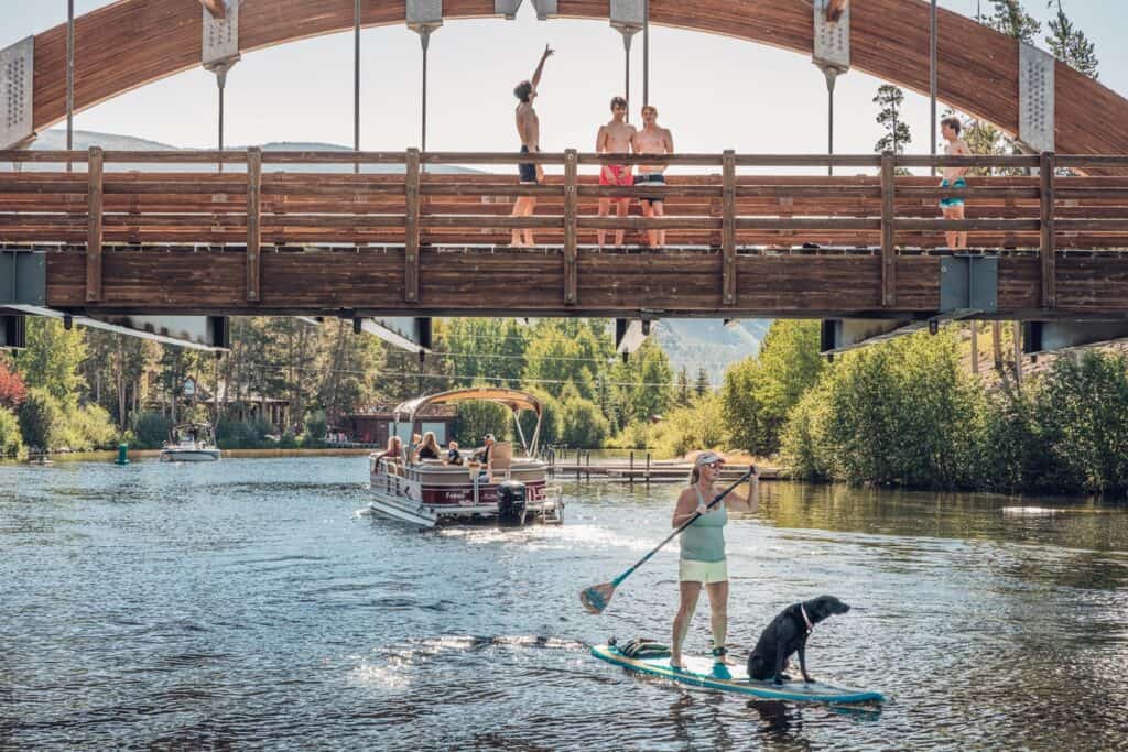 Paddleboarder and dog on Grand Lake with a boat and people on a wooden bridge in the background.