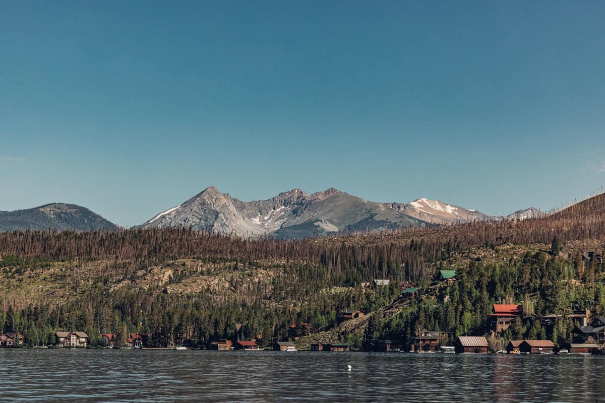 A tranquil lake is seen in the foreground with a hilly landscape dotted with rustic cabins and a backdrop of snow-capped mountains under a clear blue sky.