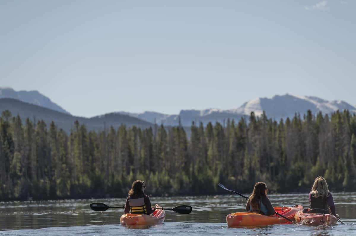 Three people kayaking on a calm lake in Grand County, Colorado, with stunning mountain peaks and dense evergreen forests in the background.