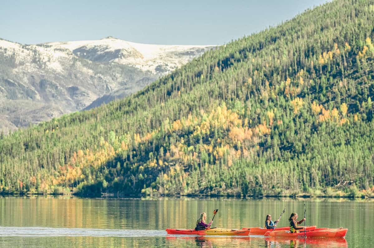 Three people kayak across Grand Lake with snowcapped mountains and golden trees in the background.