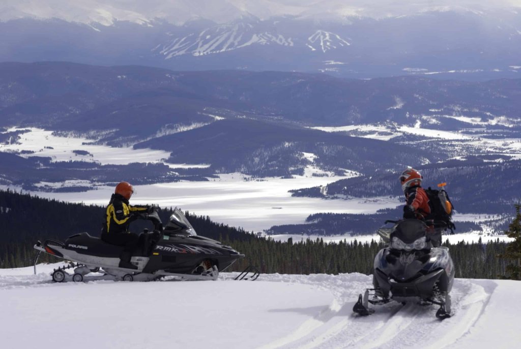 Two snowmobilers on a snowy mountain ridge overlooking a vast, snow-covered valley and distant mountains in Grand County Colorado.