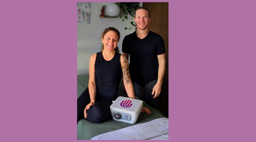 A smiling couple sits on a bed with a white device featuring a purple logo in Grand County Colorado.