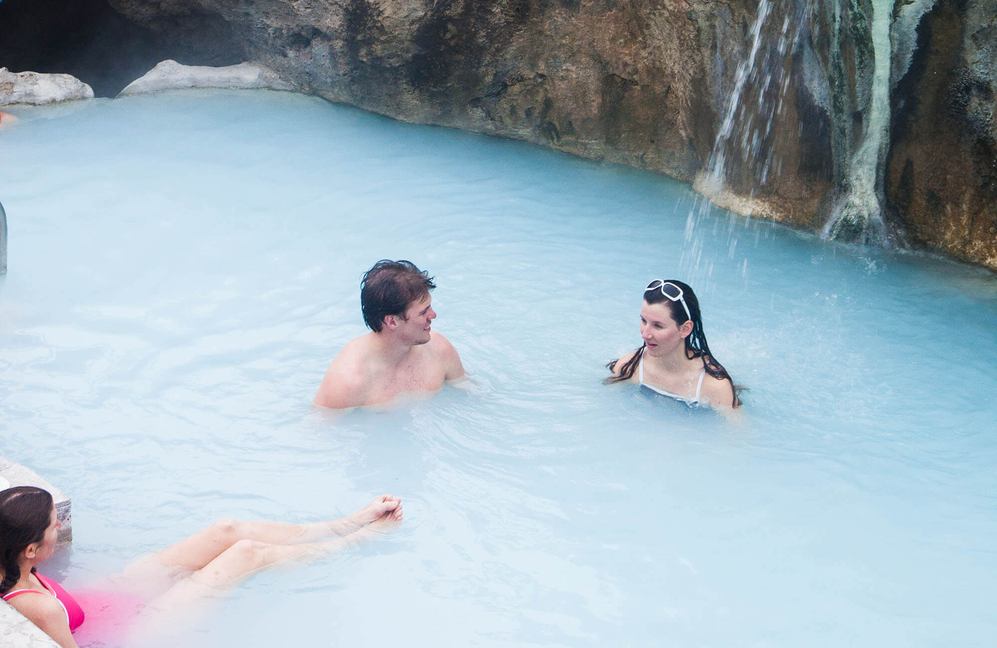 Three people relax in a serene blue hot spring pool surrounded by rocky cliffs, with a waterfall cascading into the water in Grand County Colorado.