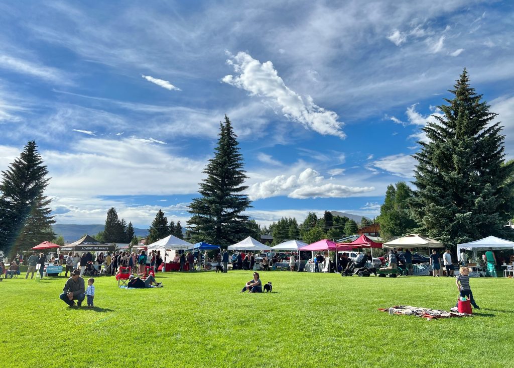 People gathered at an outdoor market in Granby with colorful vendor tents, pine trees, and blue skies in the background.