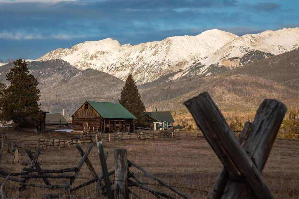 A barn in the middle of a field with snow capped mountains in the background.
