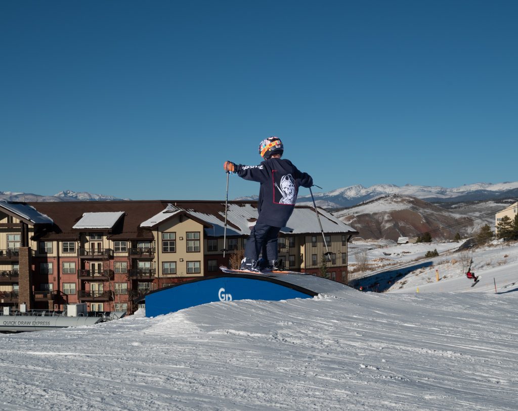 A young skier, wearing a helmet and goggles, stands poised on a snowy ramp, ready to launch into the air in Grand County Colorado.