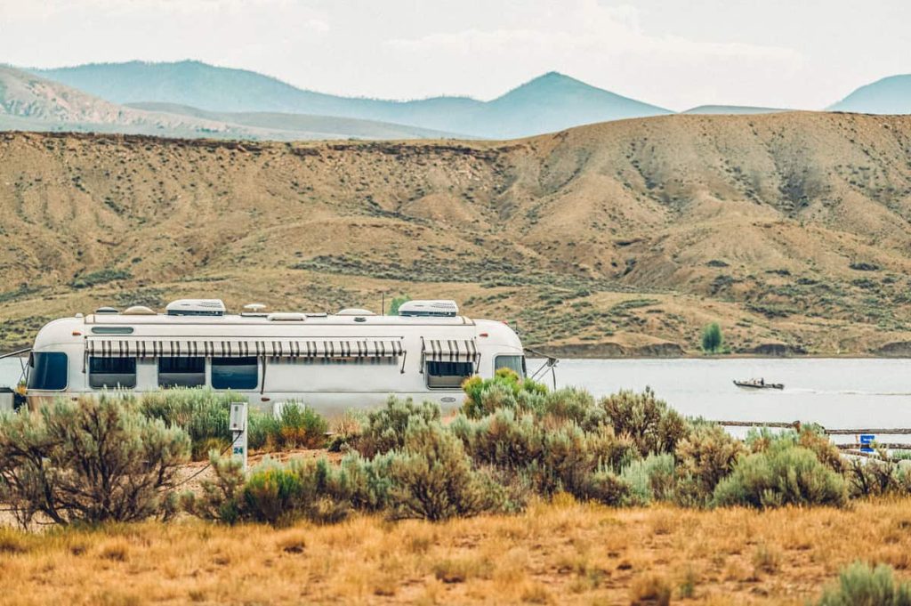 A silver Airstream trailer is parked in a desert landscape near a calm lake with mountains in the distance in Grand County Colorado.