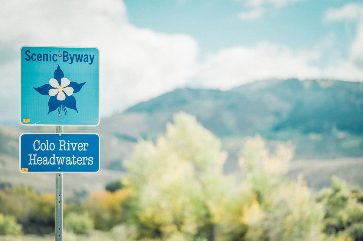 A sign reads "Scenic Byway Colo River Headwaters" with green plants and mountains in the background.