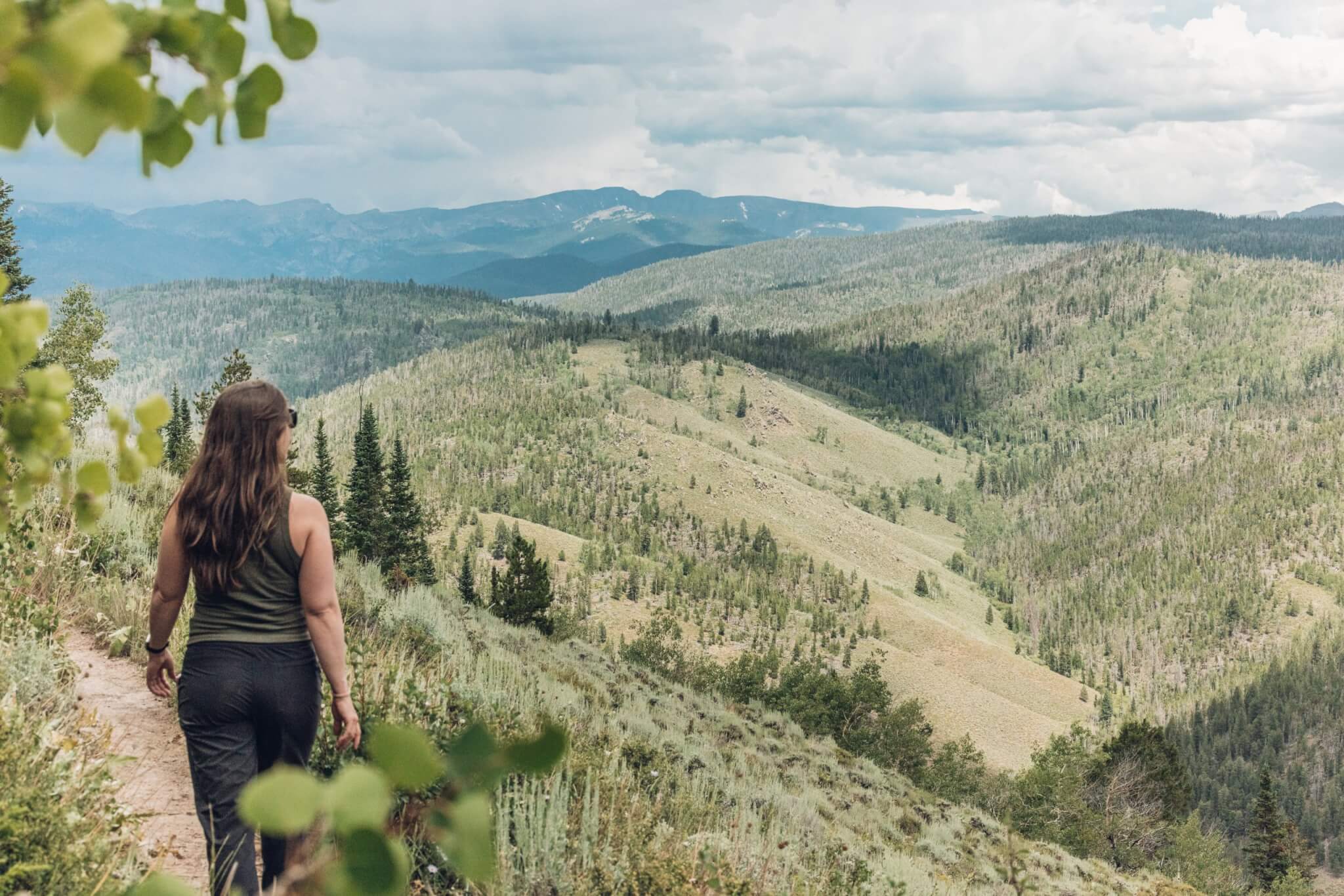 A woman in a green tank top and black pants walks a dirt path through a lush, green mountain landscape in Grand County Colorado.