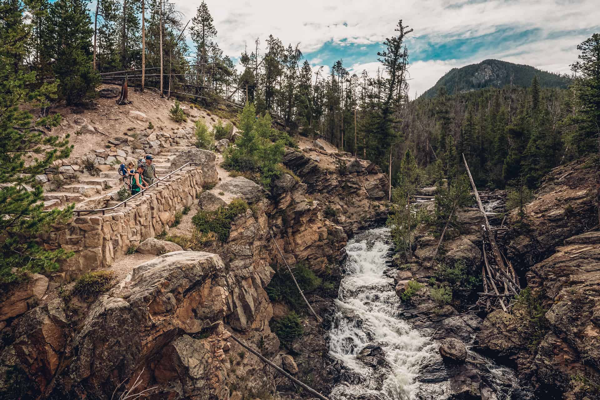 Hikers walking along a scenic stone path overlooking a rushing waterfall in Rocky Mountain National Park, surrounded by lush forest and rugged cliffs.