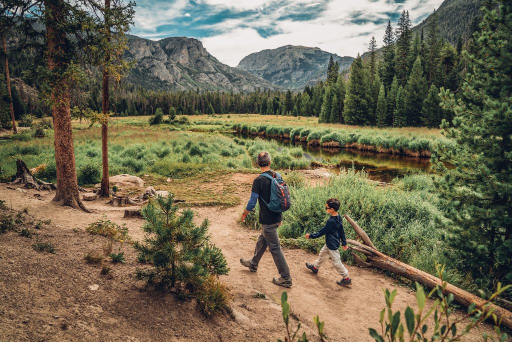 Man and child hiking along a dirt trail with grassy meadows and mountain peaks in Rocky Mountain National Park.
