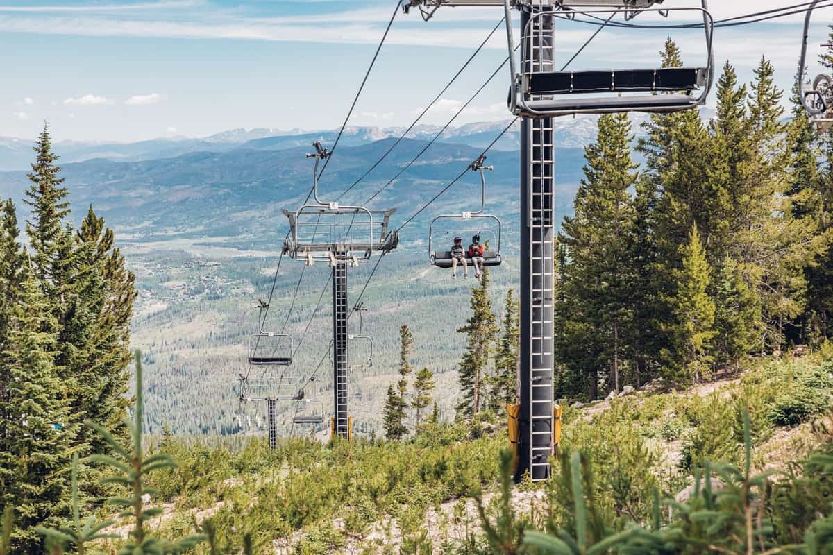 Two people sit on a ski lift, wearing bike helmets in Winter Park, with the Continental Divide in the background.