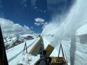 A snowplow clears thick snow from a mountainous road under a bright blue sky with scattered clouds. Snow is being thrown up to the side as the plow progresses.