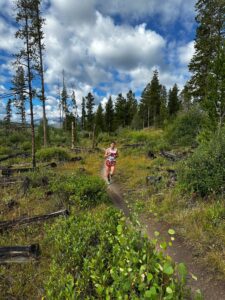 A person in running gear jogs along a narrow dirt trail through a forested area under a partly cloudy sky, surrounded by logs and green vegetation.