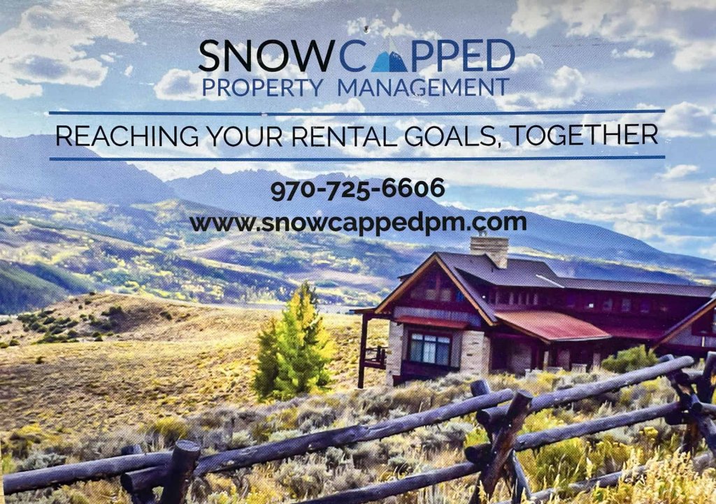 A rustic mountain cabin with a red roof is nestled in a grassy field with a wooden fence in Grand County Colorado.