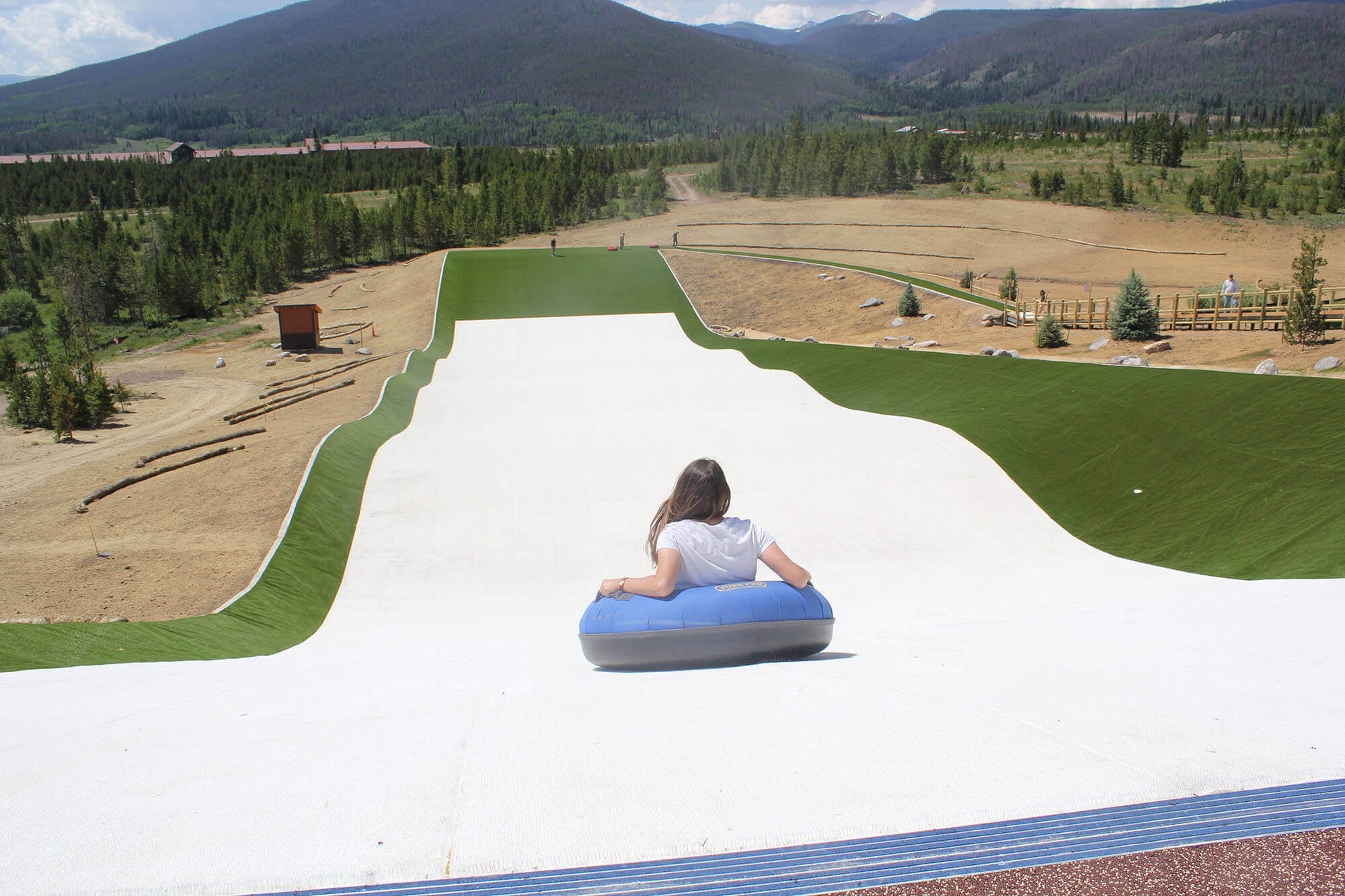 A woman in a blue inner tube glides down a long, white water slide that winds through a grassy landscape with mountains in the background at Snow Mountain Ranch in Grand County Colorado.