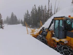 A snowplow clears snow from a road surrounded by trees in a snowy, forested area. Snow is being pushed to the side by the plow's blade.
