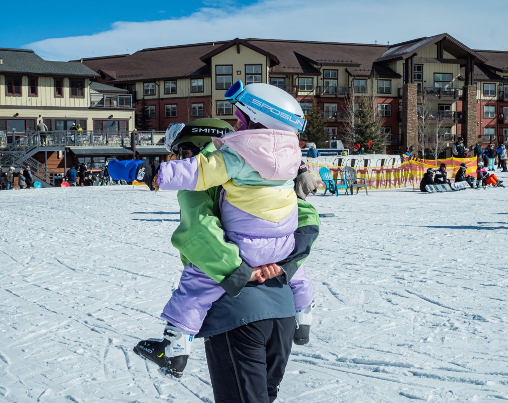 A parent carries a child dressed in colorful winter gear on their back in a snowy resort area in Grand County Colorado.