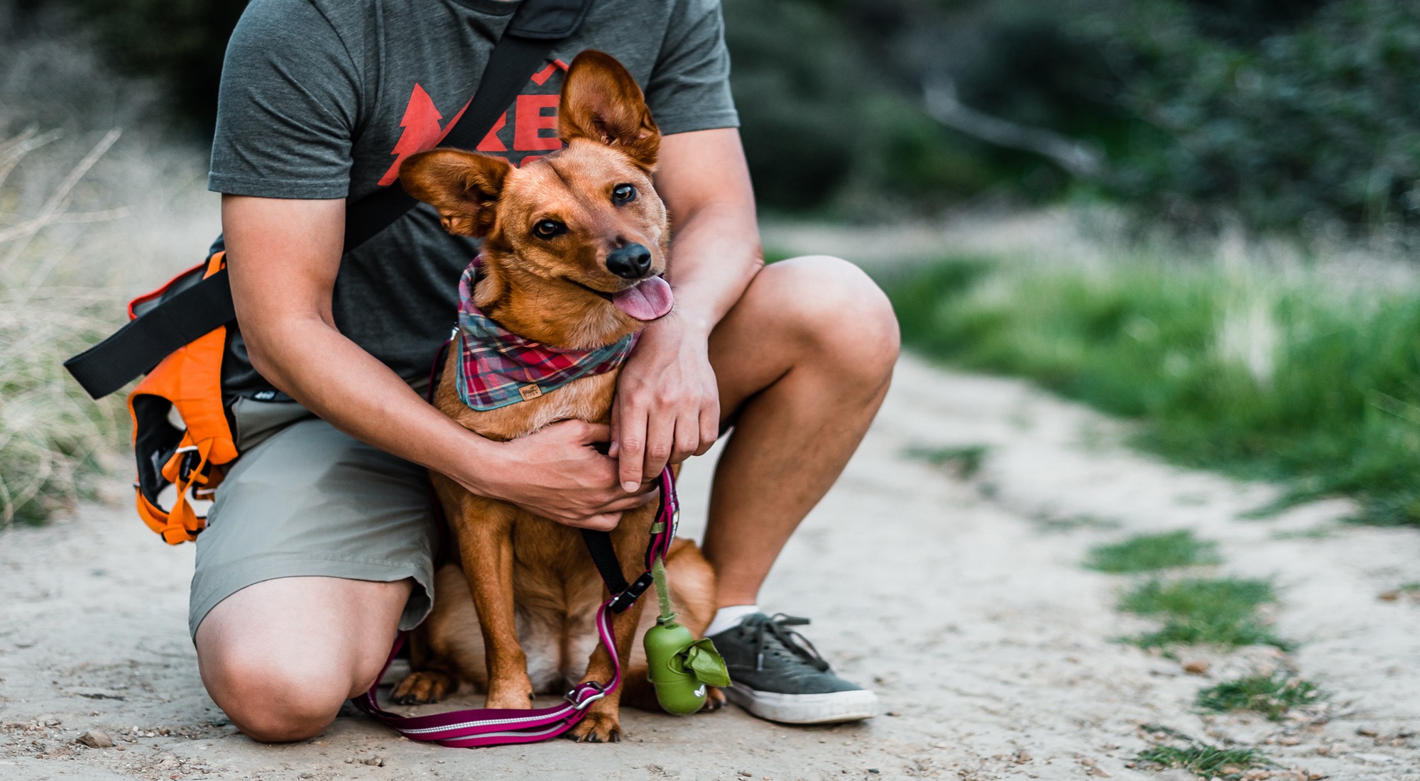 A man sits on a dirt path, hugging a dog wearing a bandana and leash in Grand County Colorado.