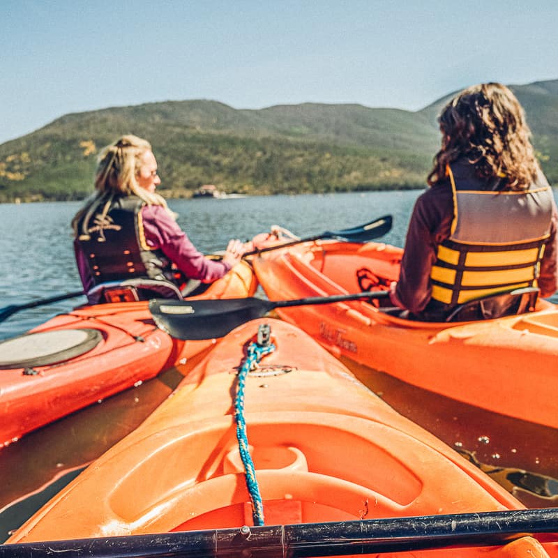 Two women kayaking on a lake with mountains in the background.