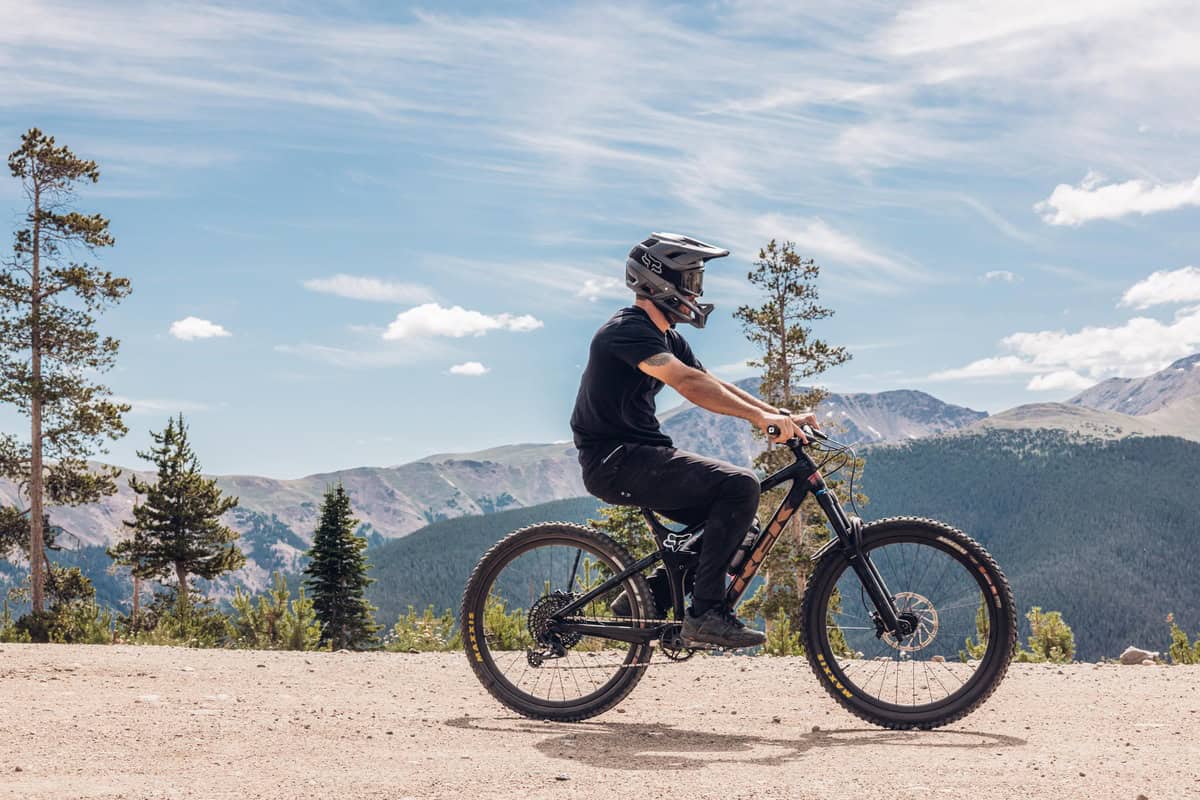 A person wearing a helmet rides a mountain bike in winter park on a trail with a mountainous landscape in the background. The sky is clear with some clouds.
