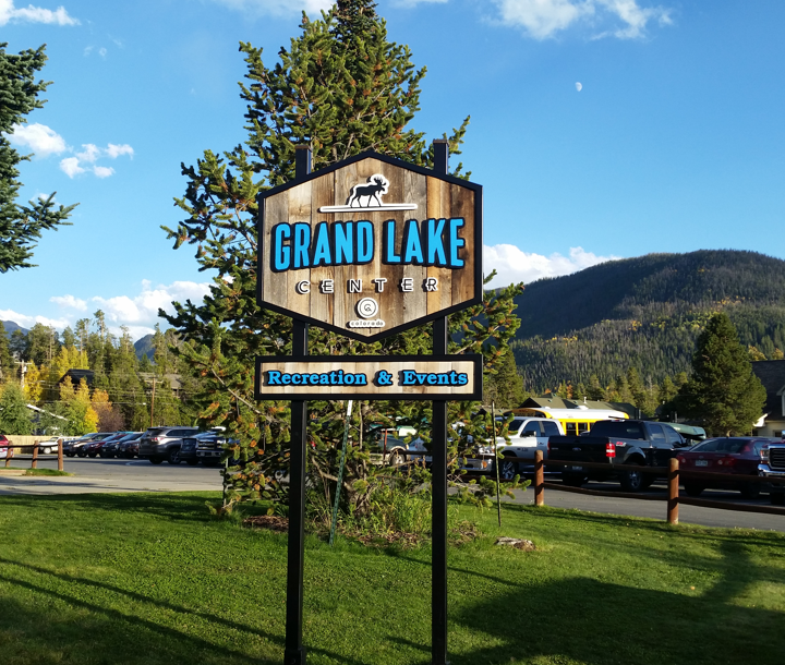 A wooden sign for the Grand Lake Center with a moose logo stands in a grassy area in Grand County Colorado.