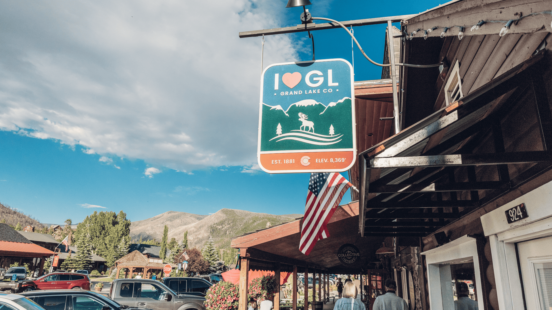 A scenic view of Grand Lake, Colorado, featuring a rustic downtown street with a "I ❤️ GL Grand Lake CO" sign, wooden storefronts, and mountain scenery in the background.