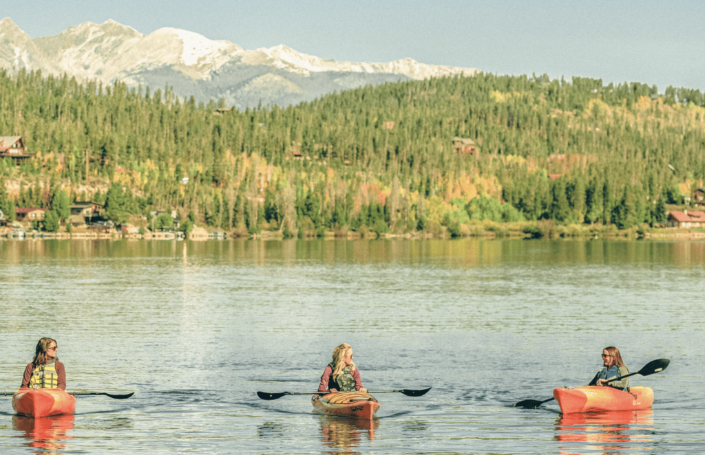 Three people kayaking on the Blue River with forested hills and snowy peaks in the background.